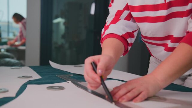 Person marks fabric with precision tool. Hands steady, focused on cutting pattern. Background shows modern office space. Ideal for fashion design, tailoring, or crafting projects
