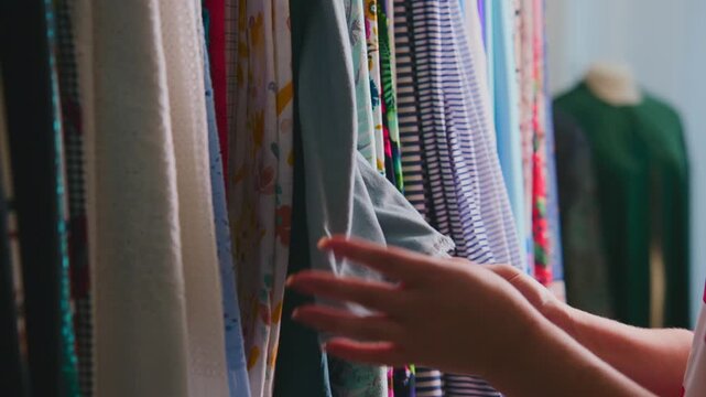 Hands gently touch fabrics on a clothing rack. Colorful textiles hang in soft focus background. Person selects garment with care and attention. Scene captures calm, focused shopping experience