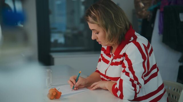 Woman in red-striped shirt writes at a table. She focuses intently on her work. Bright, natural light illuminates her workspace. Ideal for scenes of productivity, creativity, or remote work