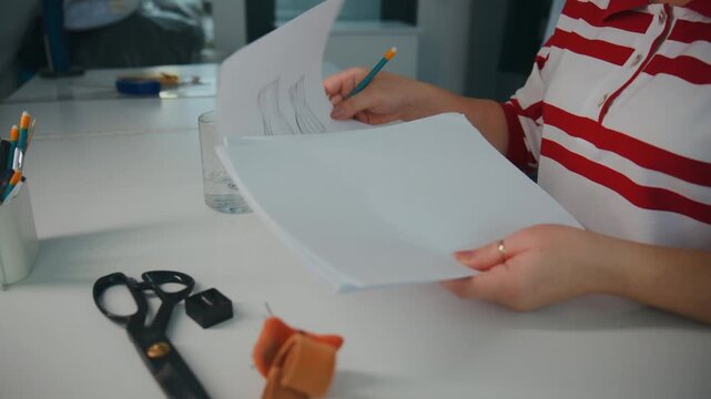 Person sketches design on white paper. Tools like scissors, pencil, and eraser nearby. Focus on creative work in modern workspace. Ideal for fashion, design, or DIY projects