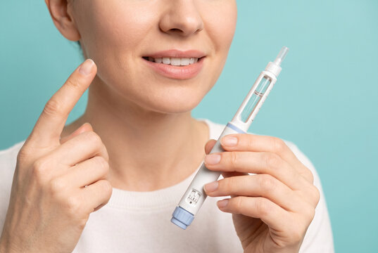 Smiling woman holding insulin pen with blue cap, pointing at cheek against turquoise background, diabetes treatment concept