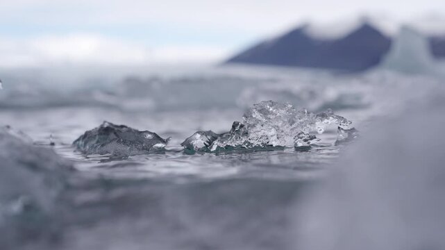Clear glacial ice chunks bobbing on rippled lagoon water with blurred icebergs and a dark mountain behind, Vatnajokull, southeast Iceland, static close up