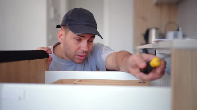 Maintenance worker uses toolbox and screwdriver in kitchen of home for repair work. Man wearing cap assembles wooden cabinet drawer with focused concentration in modern domestic interior.