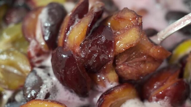 A metal spoon stirring hot halved plums and prunes boiling with pink foam during a static close up shot of cooking homemade jam in slow motion with selective focus.