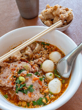 Thai spicy tom yum noodle soup with pork slices, minced pork, fish balls, crispy pork rinds, peanuts and herbs in a white bowl with chopsticks and spoon, street food lunch