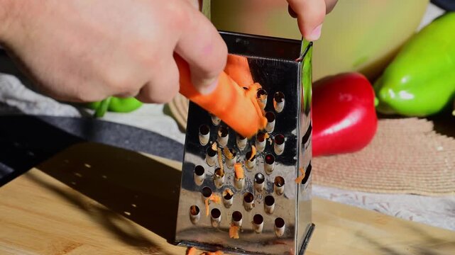 a person using a stainless steel box grater to shred a fresh orange carrot on a wooden cutting board in bright natural light
