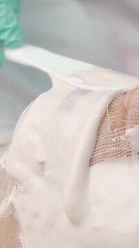 close-up motion of creamy white facial mask being applied with spatula over mesh cloth covering nose and cheek