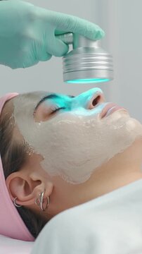woman receiving facial treatment with thick clay mask while handheld device emits blue light over closed eye,.