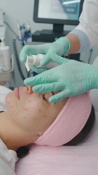 young woman with closed eyes receives facial treatment for acne in modern clinic as esthetician wearing greens