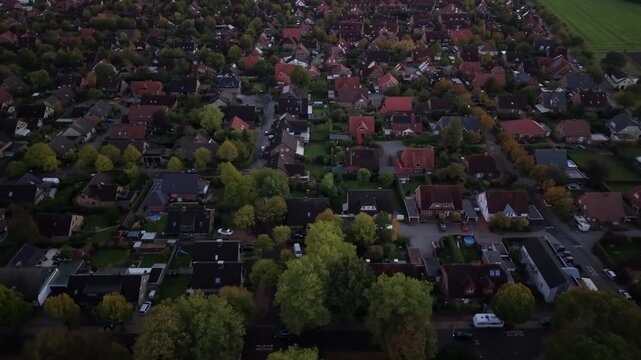 Colorful autumn trees frame quiet German neighborhood of single family homes, red rooftops and winding streets spreading through dense residential town below the canopy. Aerial drone top down shot