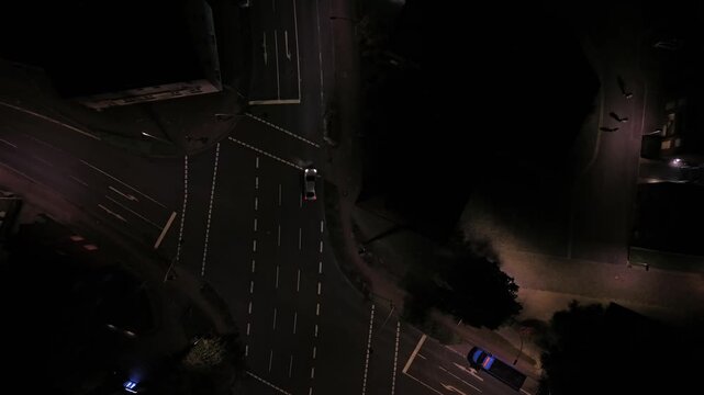 White car drives along empty multi-lane road at night, parked van and vacant parking spaces sitting beneath distant streetlamp glow across quiet American town street. Aerial drone top down