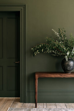 Slim wooden console table with decorative vase and greenery against olive green wall in entryway
