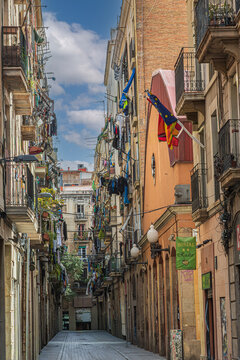 Barcelona, Spain - April 15, 2024: Narrow pedestrian street lined with historic buildings, wrought-iron balconies, hanging laundry, Catalan and European flags.