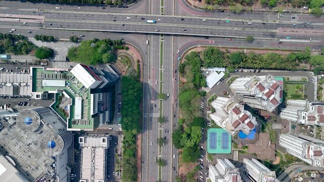 Jakarta, Indonesia - 29 September 2026: Aerial view of a busy multi-lane road separating modern high-rise apartment complexes, tennis courts, and swimming pools surrounded by green trees.