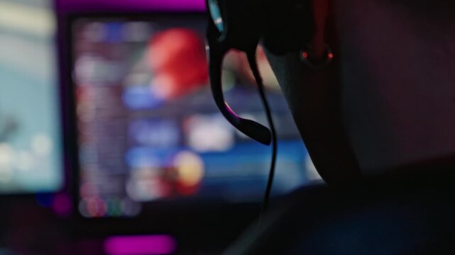 Close up over the shoulder shot of a male gamer wearing a headset and typing on an RGB backlit keyboard in a dark room. Ideal for esports, technology and lifestyle content.