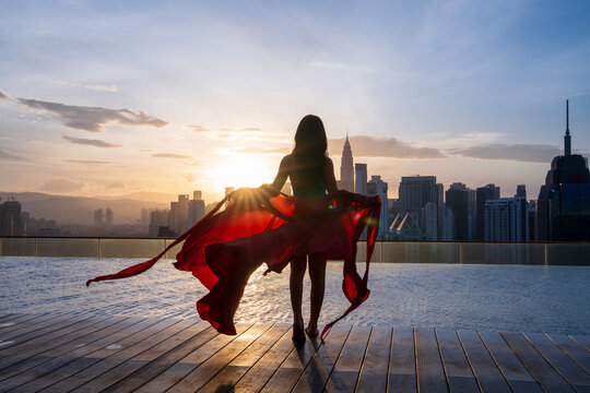 View from behind of a woman holding a flowing red fabric by an infinity pool on a rooftop, with the cityscape of Kuala Lumpur as a backdrop during a stunning sunrise in Malaysia.