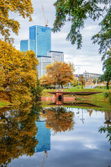 Scenic urban park in autumn with golden trees and a modern skyscraper reflecting in a calm pond surface on a cloudy day with copy space ©  Tsvyk L.