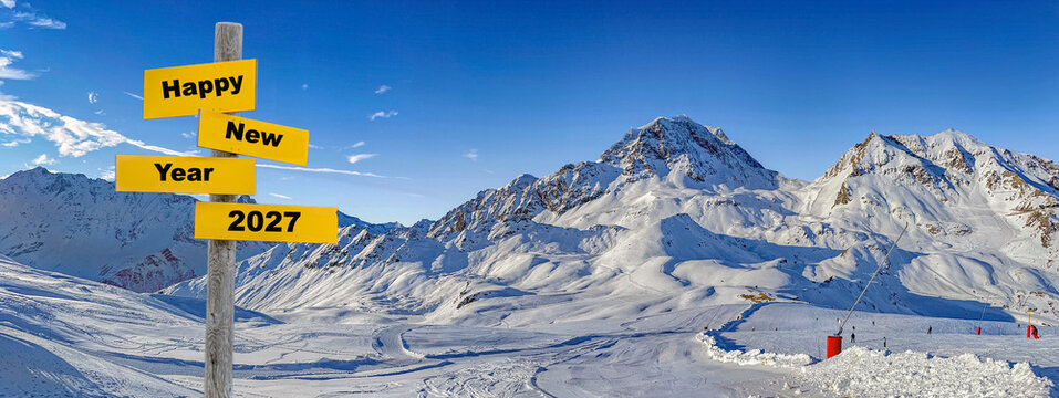 2027 Happy new year writing on yellow sign post in front of panorama on ski resort  with snowy mountain range and ski slope under blue sky