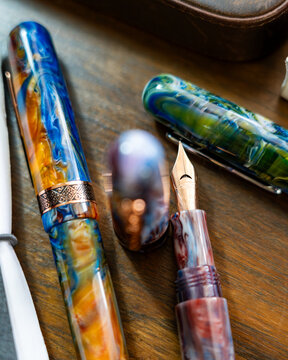 Set of luxury colorful acrylic fountain pens next to a vintage pocket watch and leather notebook on a wooden desk.