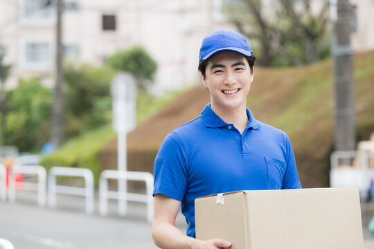 道路のあるマンションなどの屋外で段ボール箱を持って荷物を運ぶ歩く配達する若い日本人男性カメラ目線