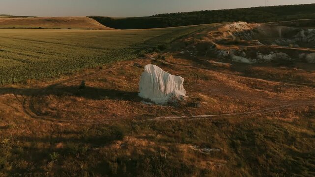 aerial pan of solitary white chalk outcrop surrounded by golden grass and agricultural fields at warm late-aft
