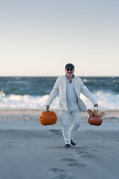 Man carrying two pumpkins along the beach at sunset. A man in a white suit walks with two pumpkins on a beach beside the sea at sunset.