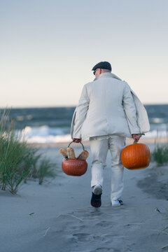 Man carries two baskets across the beach at dusk. A man in a white suit walks across a sandy beach carrying a basket and a pumpkin at dusk.