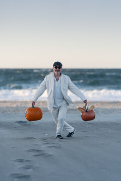 Man carrying two pumpkins across a beach at sunset. A man in a suit carries two pumpkins along a beach by the sea at sunset.