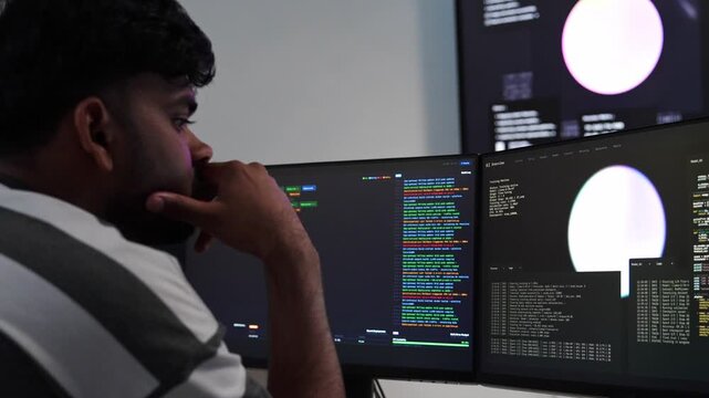 Focused Indian software developer analyzing code and artificial intelligence training data on multiple computer monitors in a dark office at night.