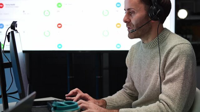 Technical support engineer wearing a headset talks and types at a computer workstation to solving customer problem in a tech office, with a large wall screen displaying call center dashboard data