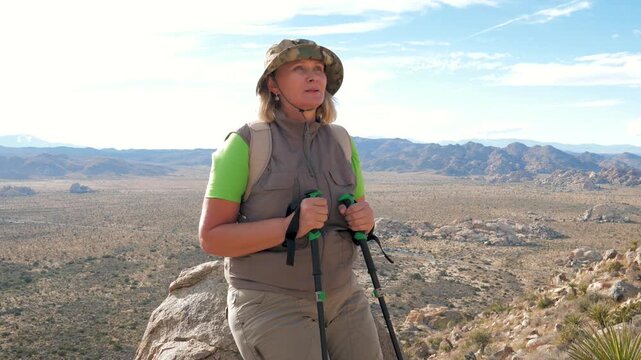 Mature female hiker holding trekking poles while standing on high rock ledge, looking at desert valley landscape during outdoor mountain trek. Female tourist is resting and recovering her breath.