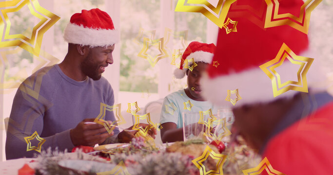 Image of stars over african american father and son in santa hats eating christmas meal
