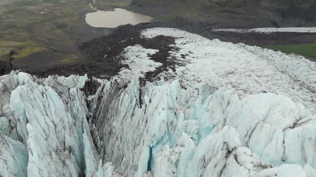 Aerial pan left over the stunning Fallj&ouml;kull glacier on Iceland, revealing rugged ice formations, a milky glacial lagoon, and vast, remote landscapes under an overcast sky.
