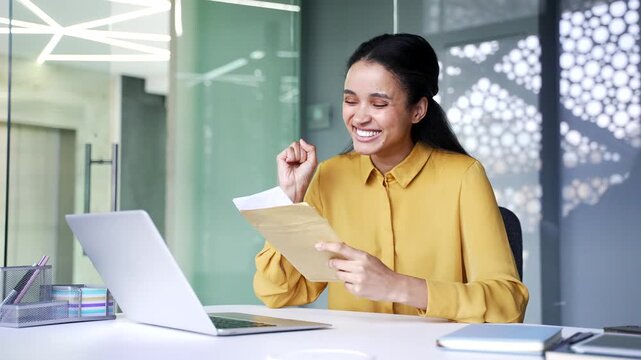 Happy young businesswoman opening envelope and reading letter with good news at office desk. Satisfied female employee looking at document and smiling after receiving positive message.
