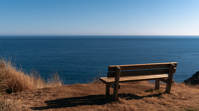A wooden park bench sits on a grassy cliff edge overlooking the blue ocean. This peaceful scene represents travel, meditation, and quiet reflection.