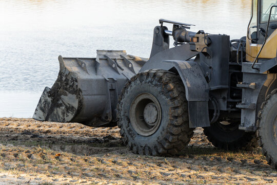 Dirty excavator bucket and tracks on sandy shore near water at construction site copy space