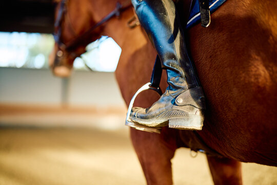 Equestrian rider wearing a tall leather boot and placing a foot in the stirrup beside a chestnut horse in an indoor riding arena
