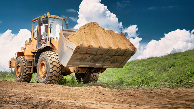 A front end loader carries soil in its bucket on a dirt track beside a grassy field during the day