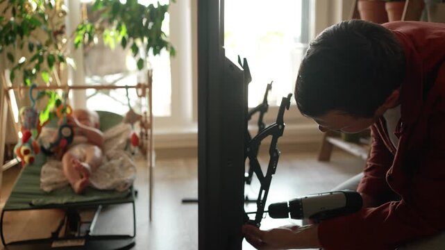Young multitasking father assembling a new television wall mount in his living room, carefully using a cordless drill while his infant son rests and plays nearby in a bouncer