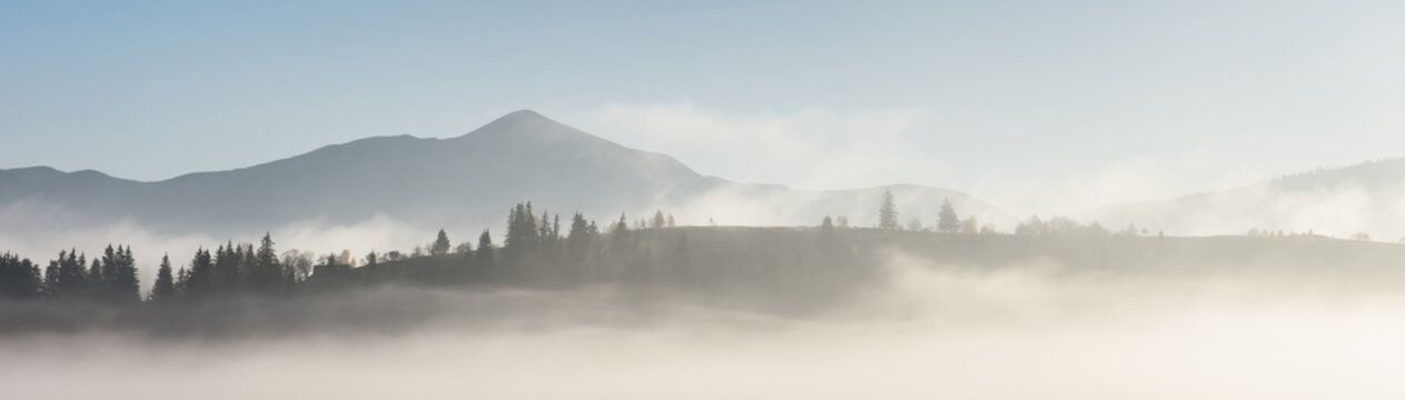 Layers of distant mountain ridges emerge through low morning fog beneath a pale sky. Soft light and muted colors create a minimalist panoramic landscape
