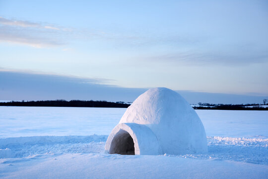 A small snow igloo stands on a frozen field beneath a pale blue winter sky. Smooth snowdrifts and a distant dark horizon create a minimalist Arctic landscape
