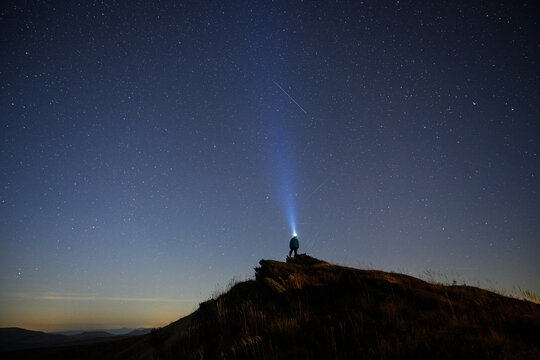 A lone man stands on a rocky hill beneath a star-filled night sky with a bright beam of light rising upward. A dark silhouette and distant horizon create a dramatic astronomical scene