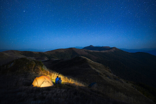 A glowing tent sits among dark mountain ridges beneath a clear starry sky. Remote wilderness and two hikers near his tent. Atmospheric night camping scene