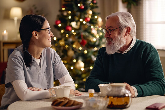 Caucasian female nurse talking with senior Caucasian man at table near Christmas tree. Image representing elderly care during holidays for geriatric healthcare and assisted living campaigns