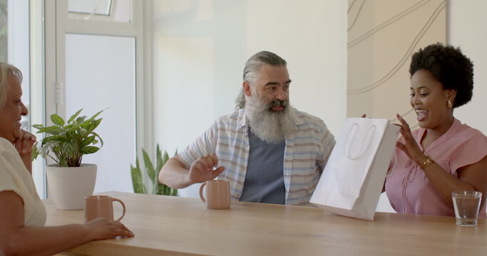 Senior couple enjoying coffee and opening gift bag at home with friend