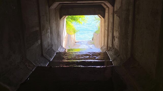 Concrete drainage tunnel carrying shallow water toward a bright lakeshore, with turquoise water and green trees framed by rough walls, creating a strong contrast between darkness and daylight