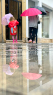 Child and adult walking through rain beneath umbrellas, reflected in wet pavement outside an urban storefront