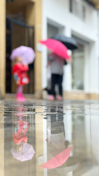 Child and adult carrying umbrellas in the rain, creating colorful puddle reflections on a wet city sidewalk