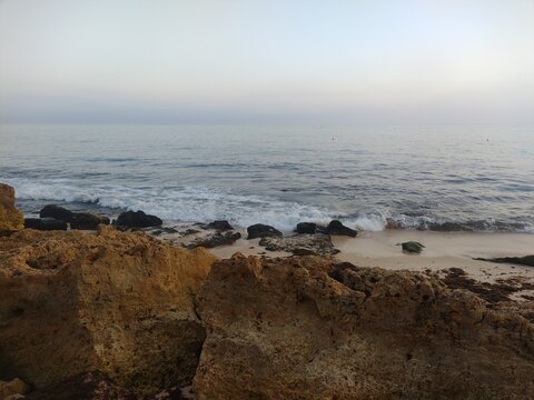 During the daytime, ocean waves roll toward a rugged, rocky shoreline beneath a soft, hazy sky, creating a calm and atmospheric coastal scene