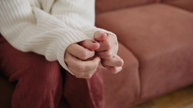 Close-up of a nervous woman fidgeting with her hands and sweater sleeves while sitting on a couch, showing signs of anxiety, stress, or emotional discomfort.